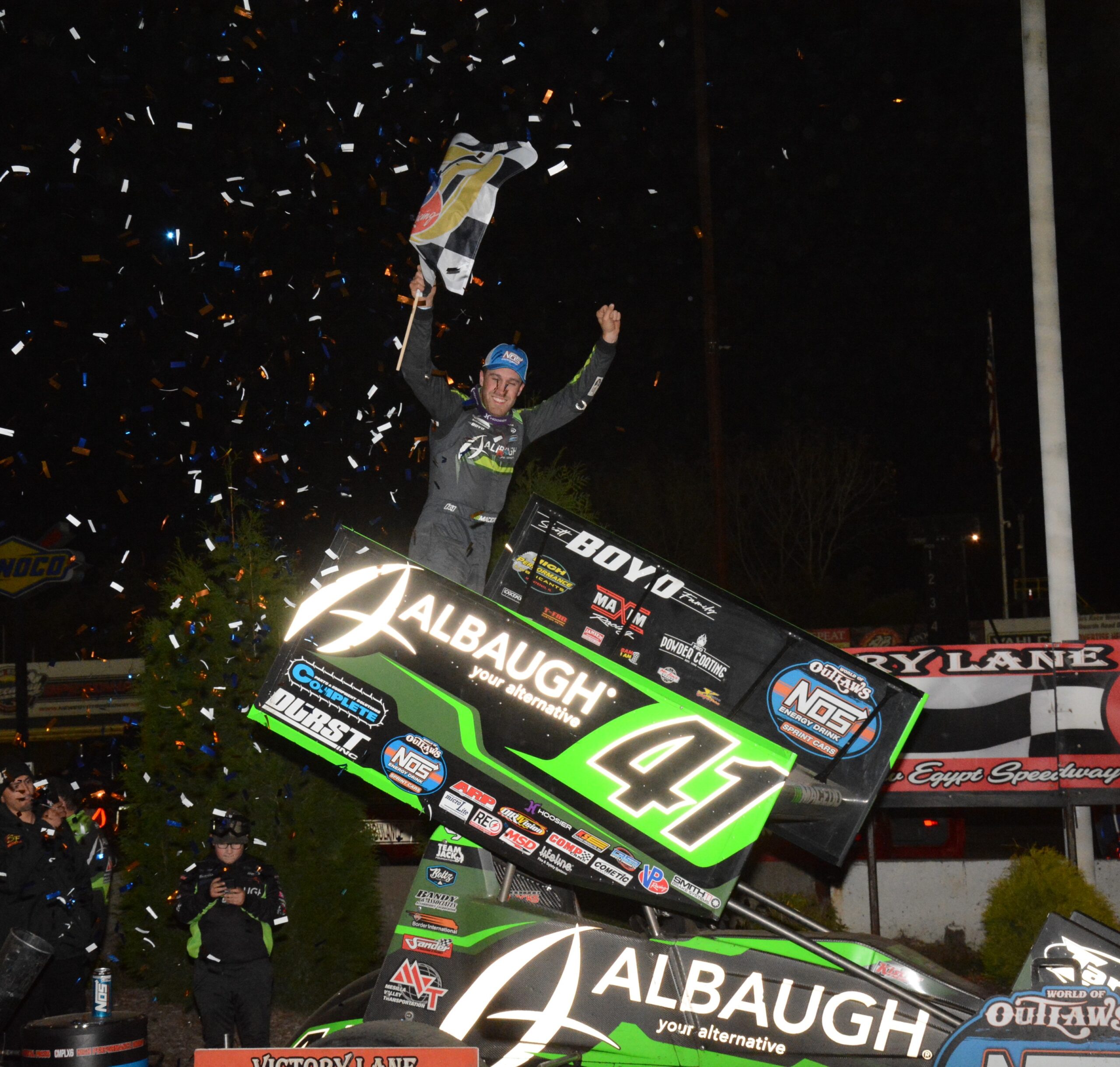 Victory lane at New Egypt Speedway — World of Outlaws winner celebrating under the confetti A winning sprint-car driver celebrates on his wing in victory lane at New Egypt Speedway, checkered flag up and confetti falling
